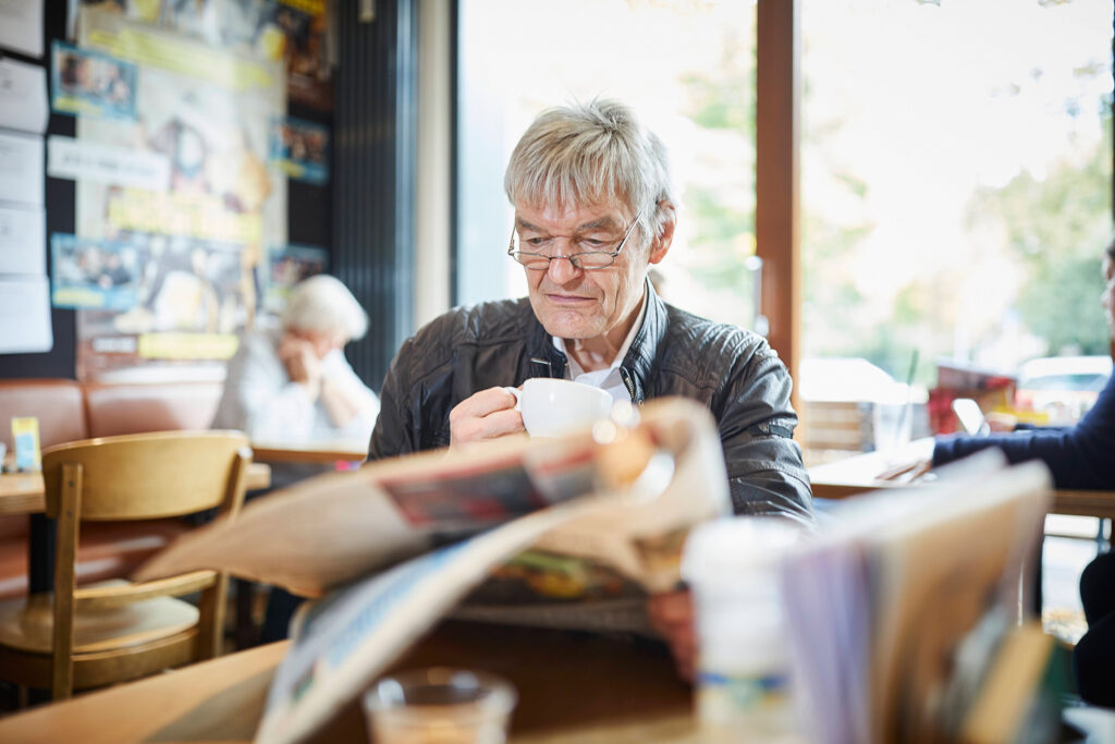 Foto: Älterer Mann sitzend im Café mit Zeitung und einer Tasse Kaffee in der Hand, Peter Umlauf, Ehrenamtler in der Sprachförderung der Kinder-, Jugend- und Familiendienste