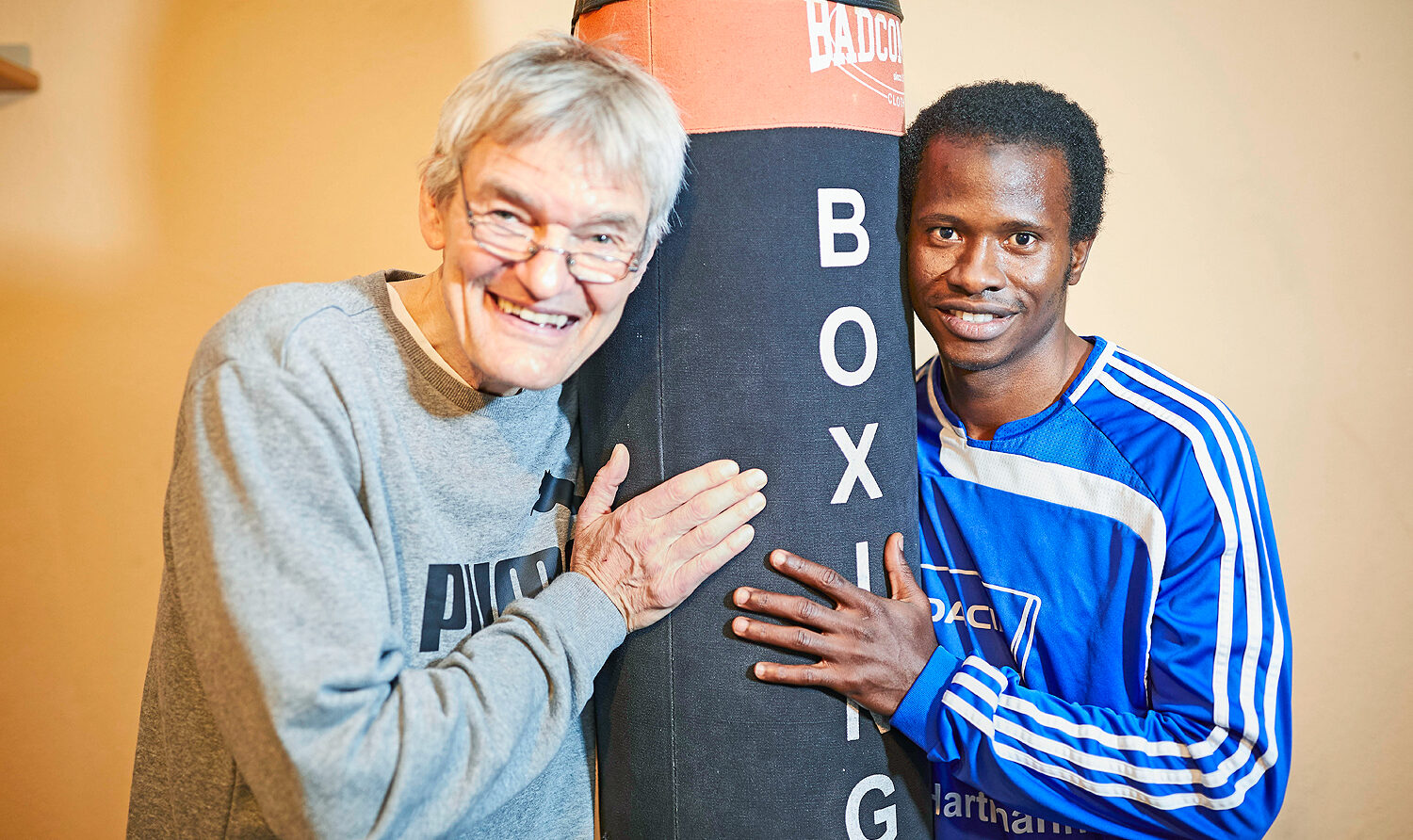 Foto: Älterer Trainer und junger Sportler mit Boxsack inder Mitte, Peter Umlauf betreut die Jugendlichen auch im Fitnessraum